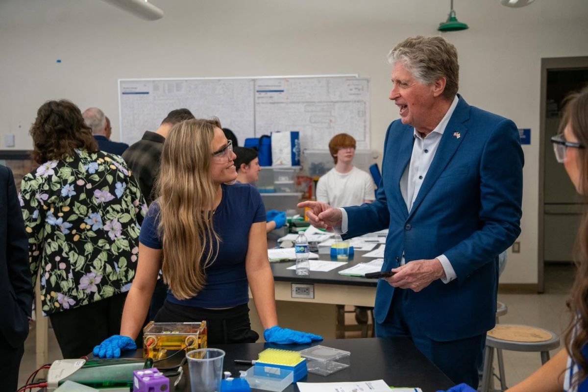 Tall older man standing in a classroom next to a female high school student wearing lab goggles and gloves